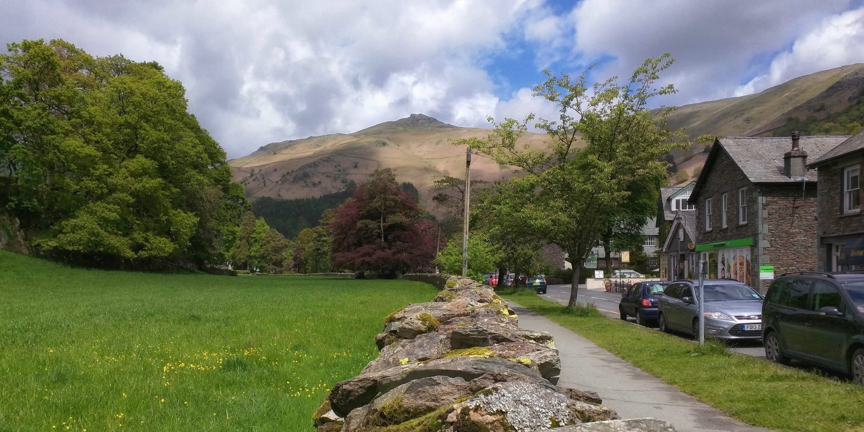 A view from a dry stone wall in the middle of Grasmere village towards the fells of Heron Pike in the distance, surrounded by old buildings and greenery